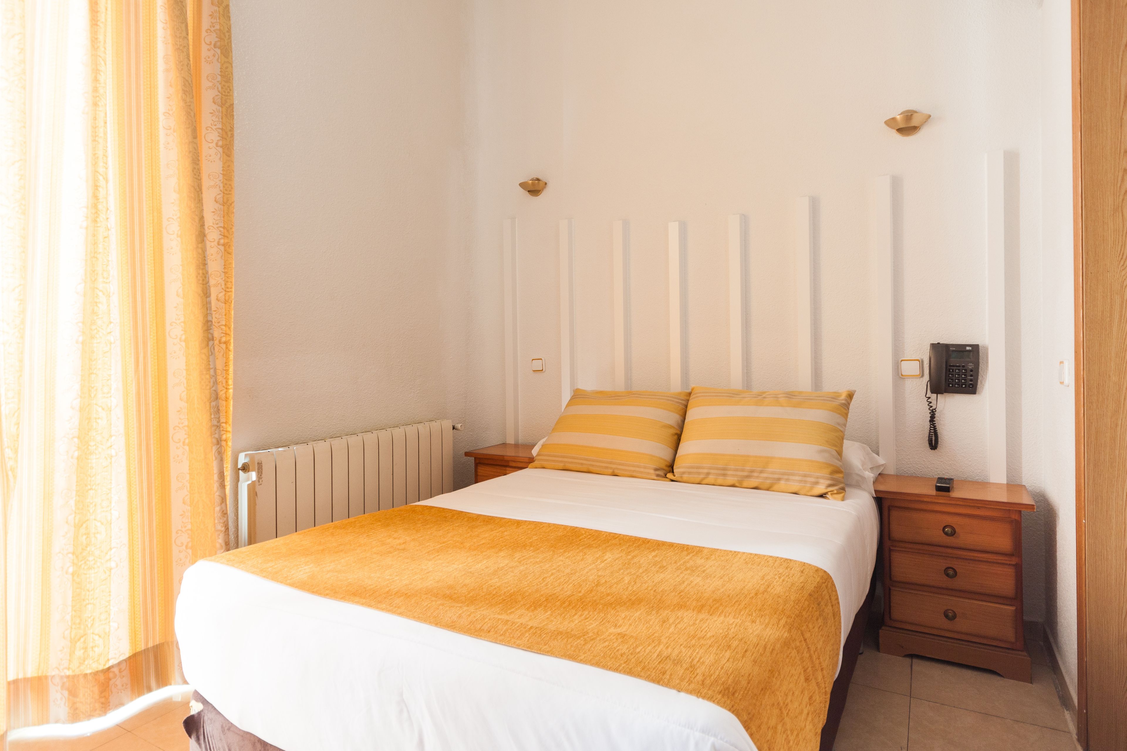 A neatly made bed with a white and orange bedspread, a wooden nightstand with a phone, and a radiator on the wall.