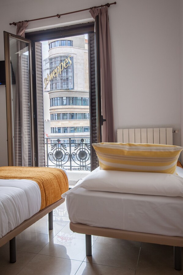 A hotel room with two beds, a view of a building with 'Schweppes' signage, and a balcony with a metal railing. - Hostal Josefina (Madrid)