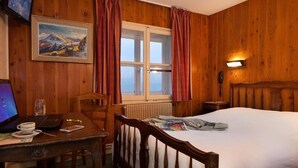 Una habitación de madera con una cama, un escritorio con un ordenador portátil, un cuadro y una ventana con cortinas.