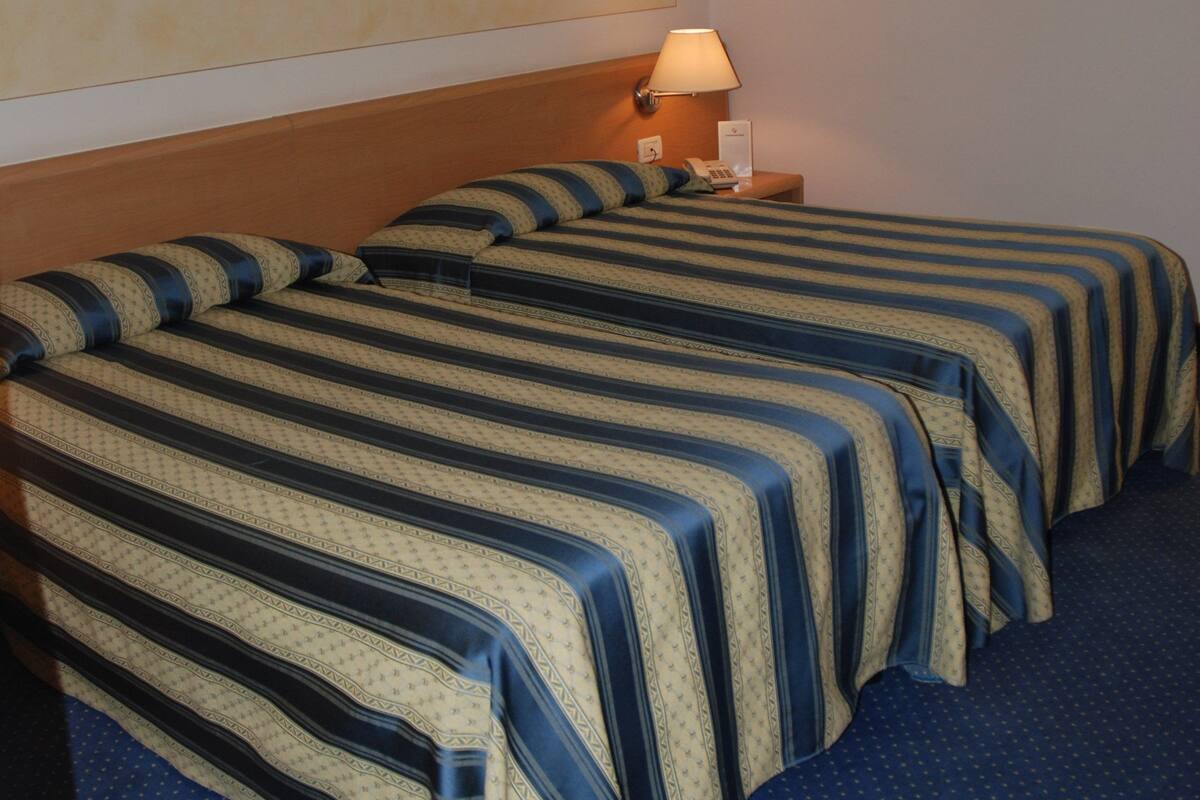 Una habitación de hotel con dos camas, una mesita de noche con lámpara y una pared lisa.