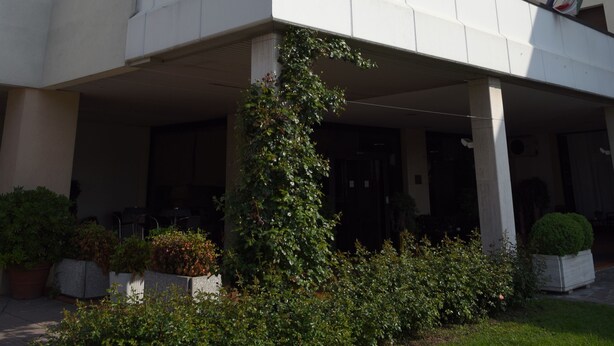 Un moderno edificio de hotel con un logotipo destacado, varios balcones y una zona de entrada cubierta con plantas en macetas.