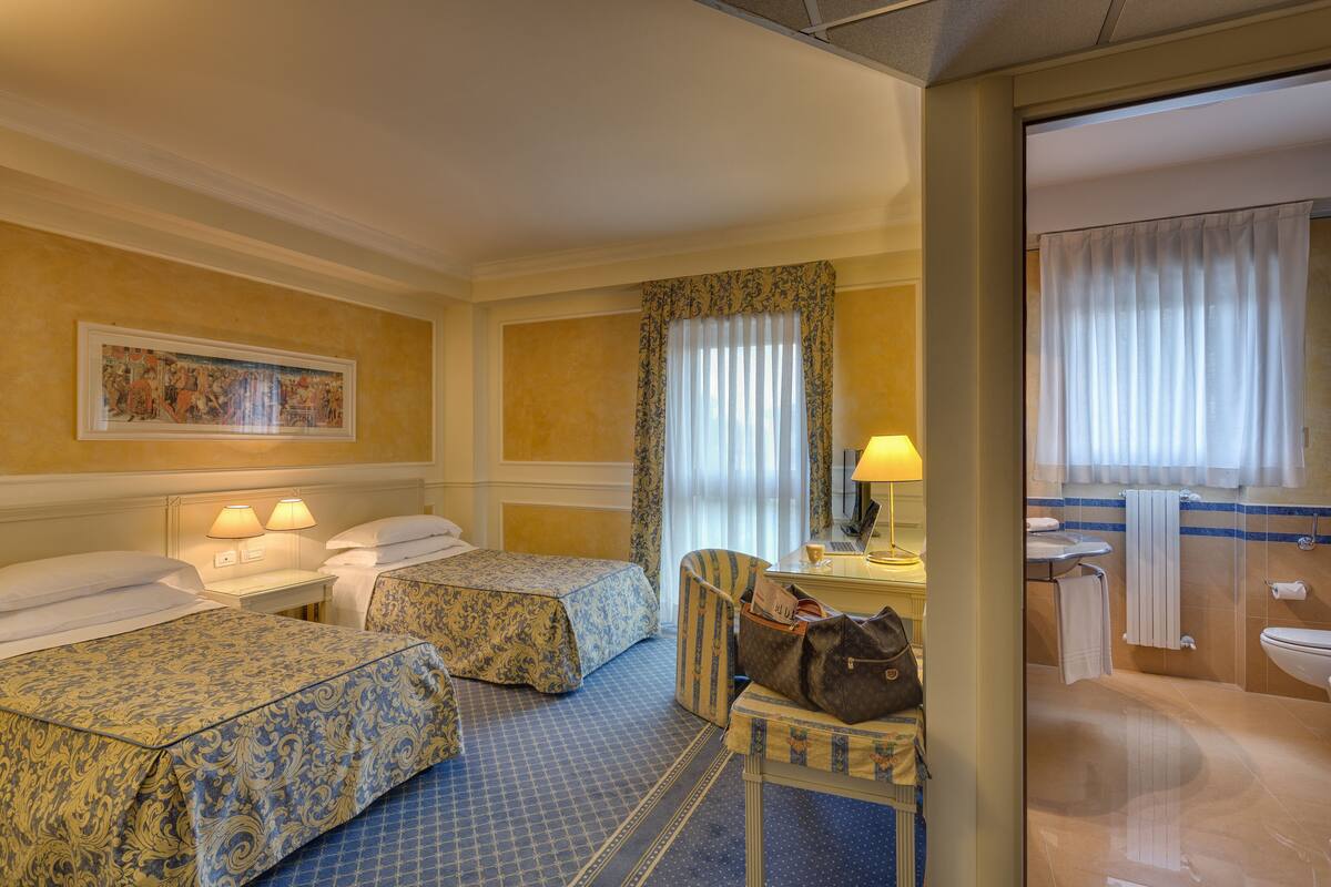 Habitación de hotel con dos camas, un escritorio con lámpara, una silla y un baño visible a través de una puerta abierta.
