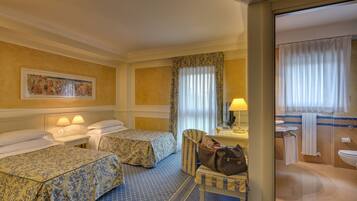 Habitación de hotel con dos camas, un escritorio con lámpara, una silla y un baño visible a través de una puerta abierta.