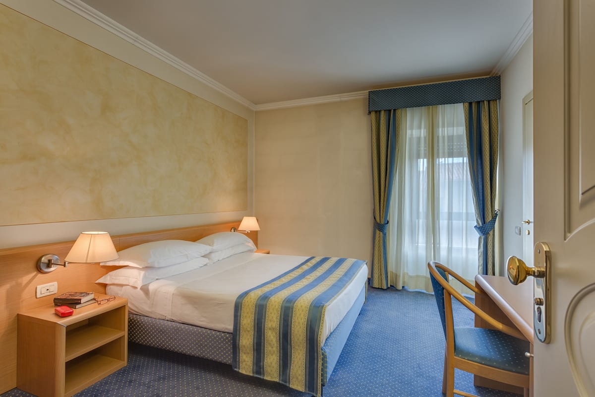 Una habitación de hotel con cama, mesita de noche, lámpara, silla y cortinas.