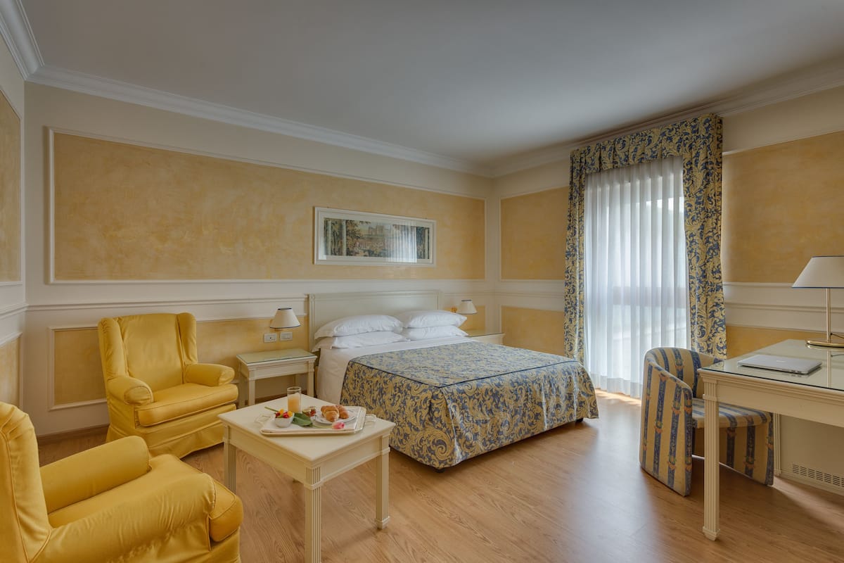 Habitación de hotel con cama, escritorio, silla, una mesita con desayuno y ventana con cortinas.
