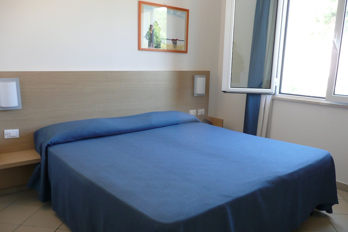 Una cama individual con colcha azul en habitación con cabecera de madera, ventana y lámpara fijada a la pared.