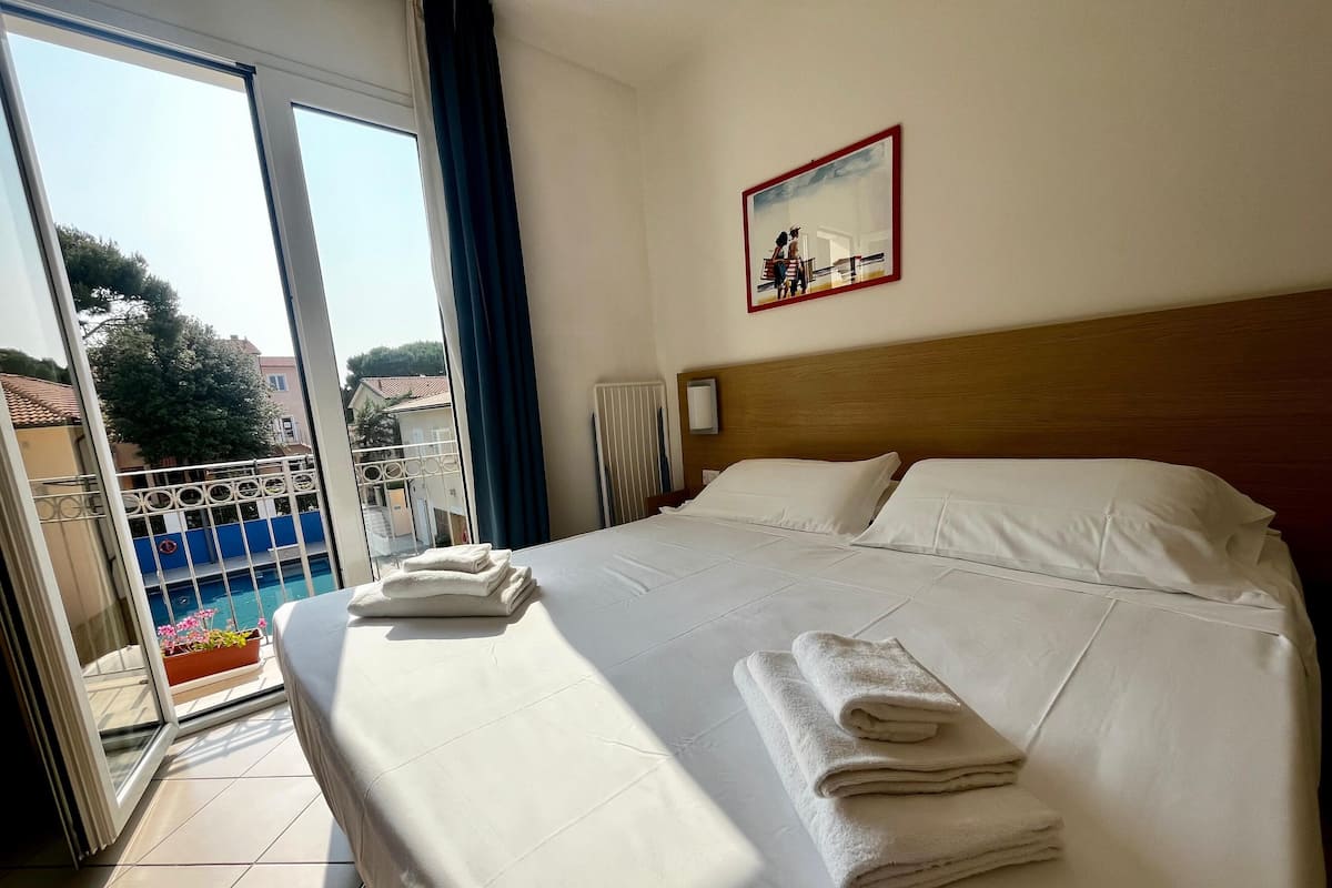 Habitación de hotel con una cama grande, dos almohadas blancas, un cabecero de madera, un cuadro en la pared y vistas a una piscina a través de una puerta de cristal.