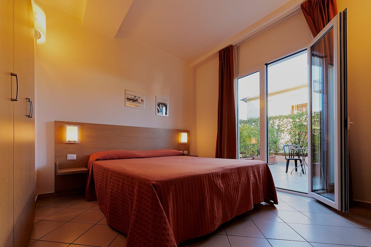 Habitación de hotel con una cama grande, dos mesitas de noche, una lámpara de pared y un ventanal corredero a un espacio exterior.