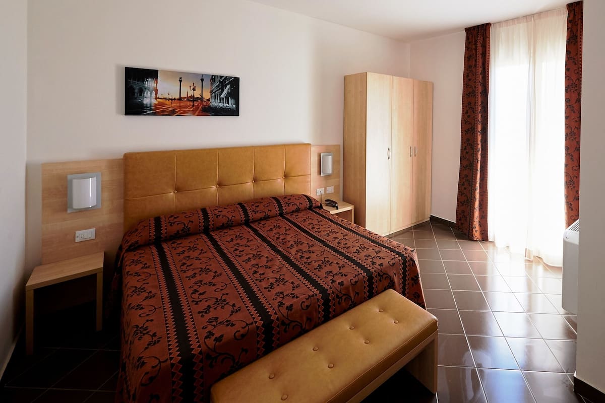Habitación de hotel con cama, banca, armario, mesita de noche y un cuadro en la pared.