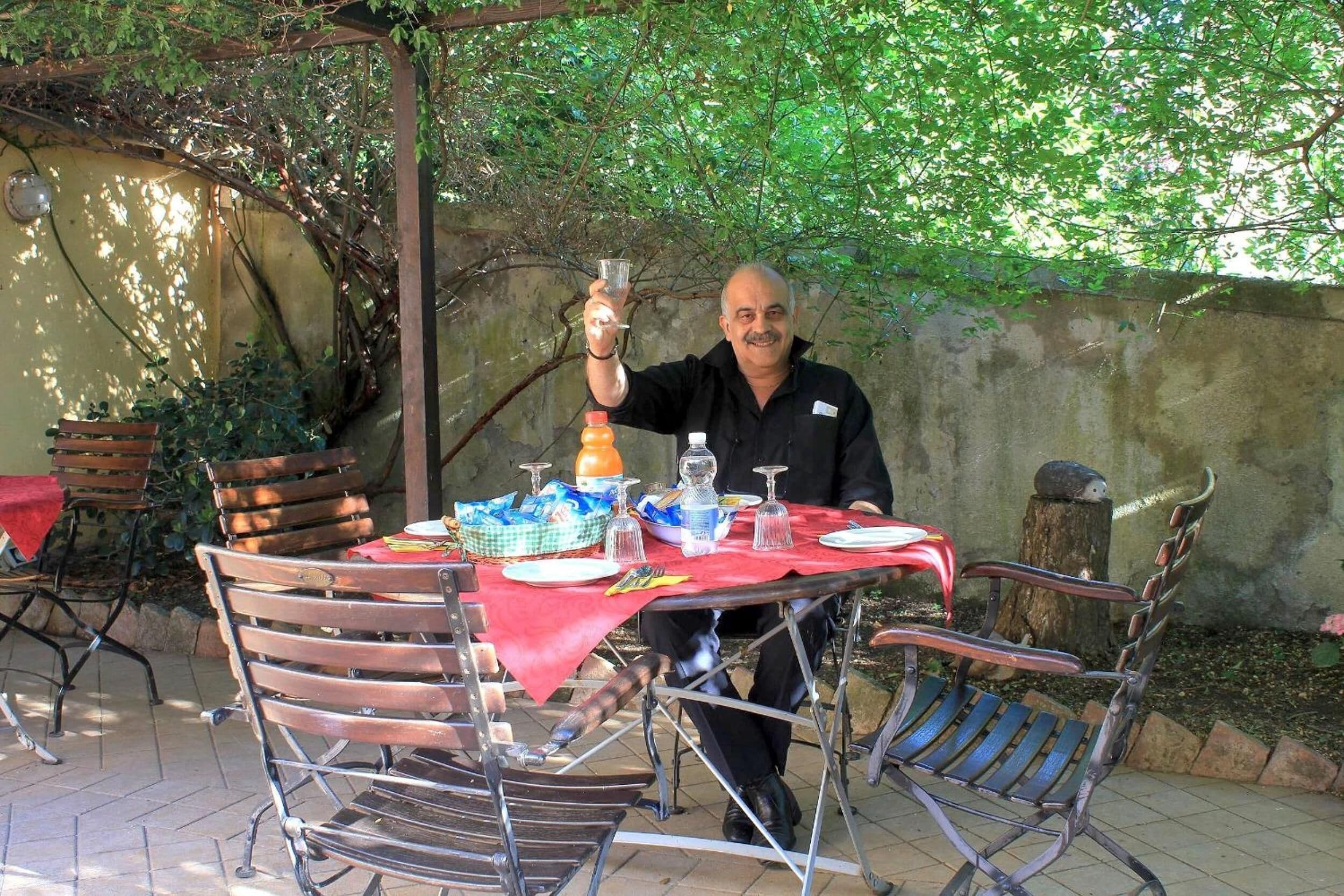 Ein Mann sitzt an einem Tisch im Freien mit rotem Tischlein, umgeben von Metallstühlen und einem Esskorb.
