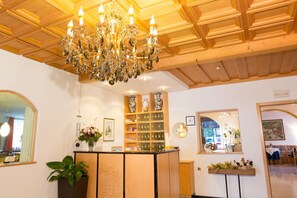 Un hall d’hôtel avec un lustre, un plafond en bois, un comptoir d’accueil, des étagères ornées d’objets décoratifs et un miroir.