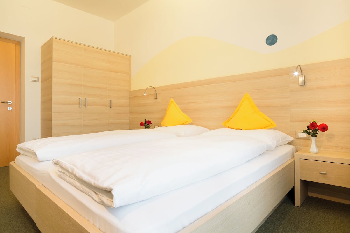 A double bed with white linens, a wooden wardrobe, and a nightstand with a vase of flowers.