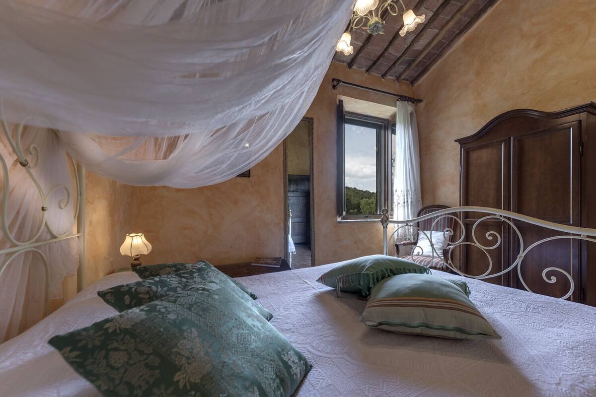 A bedroom with a canopy bed, a wooden wardrobe, and a window with a view of greenery.