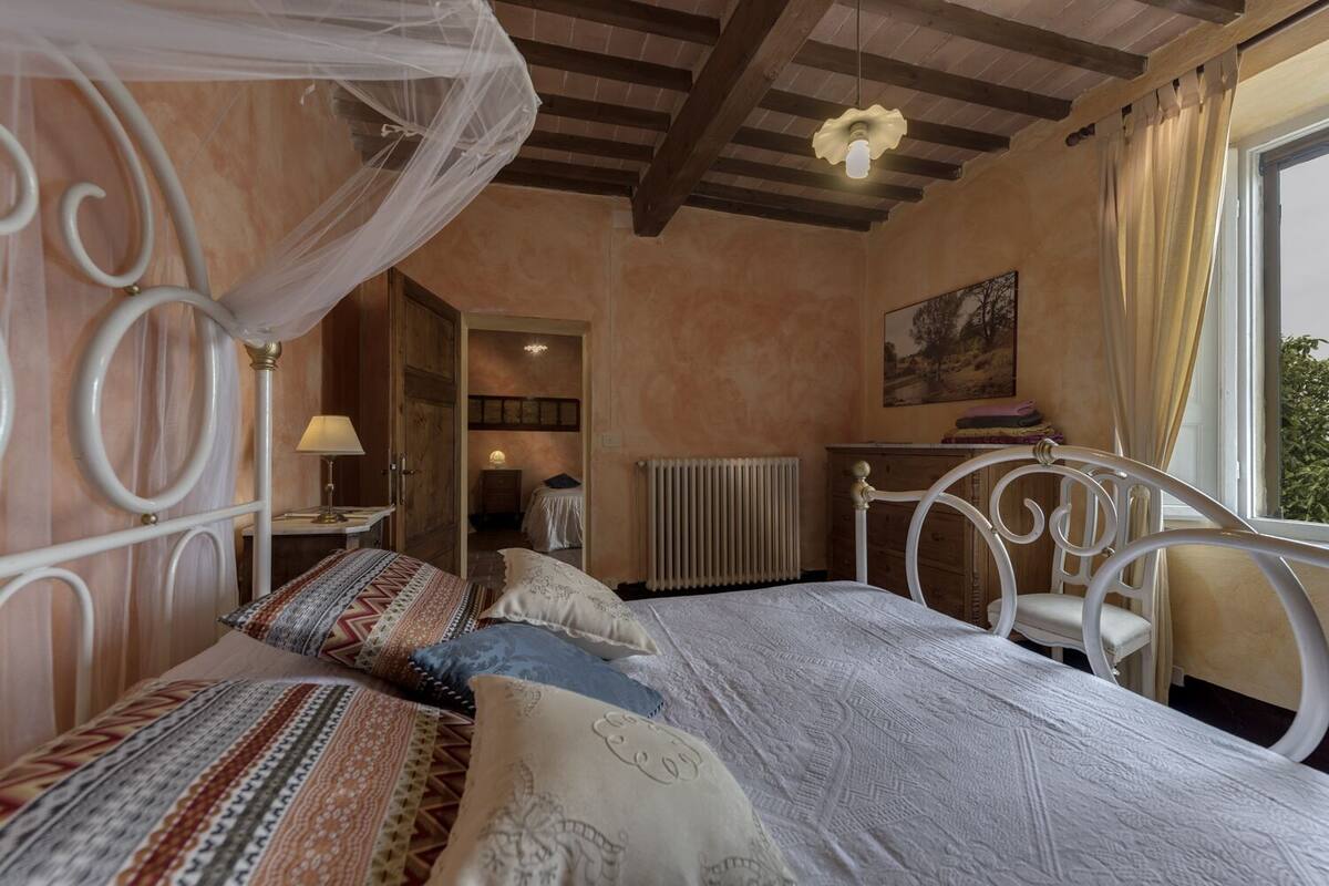 A bedroom with a bed, a nightstand, a chair, a window with curtains, a radiator, and a door leading to another room.