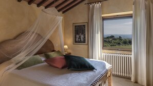 A bedroom with a bed, a nightstand, a bedside lamp, a window with curtains, and a view of the landscape. - Agriturismo La Palazzina (Murlo)