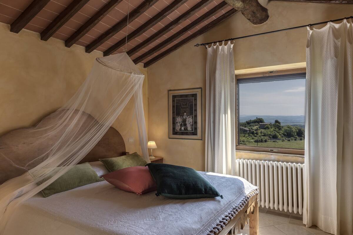 A bedroom with a bed, a nightstand, a bedside lamp, a window with curtains, and a view of the landscape.
