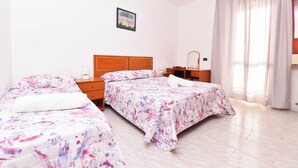 Una habitación de hotel con dos camas, un cabecero de madera, una mesita de noche con una lámpara, un escritorio pequeño y una ventana con cortinas.