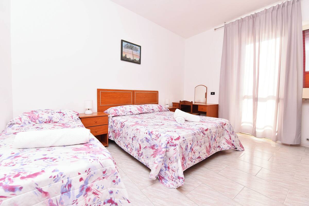 A hotel room with two beds, a wooden headboard, a bedside table with a lamp, a small desk, and a window with curtains.