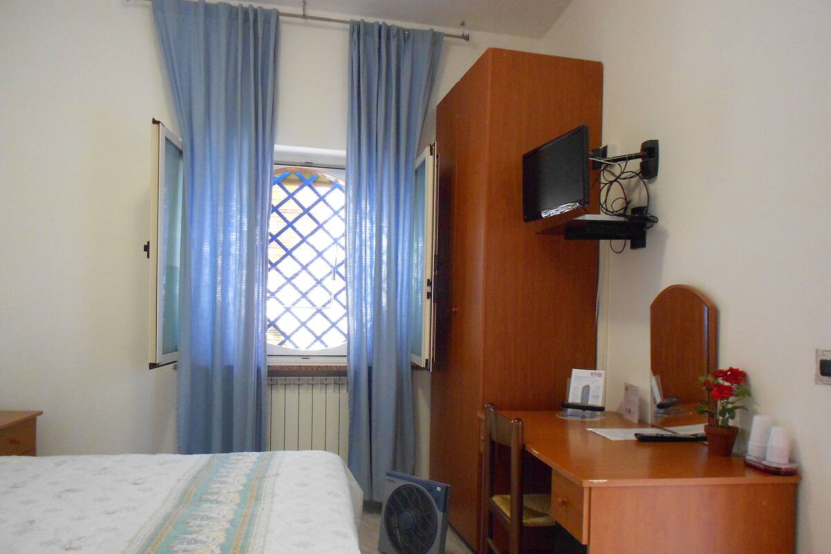 A bedroom with a bed, a desk, a chair, a TV, and a window with curtains.