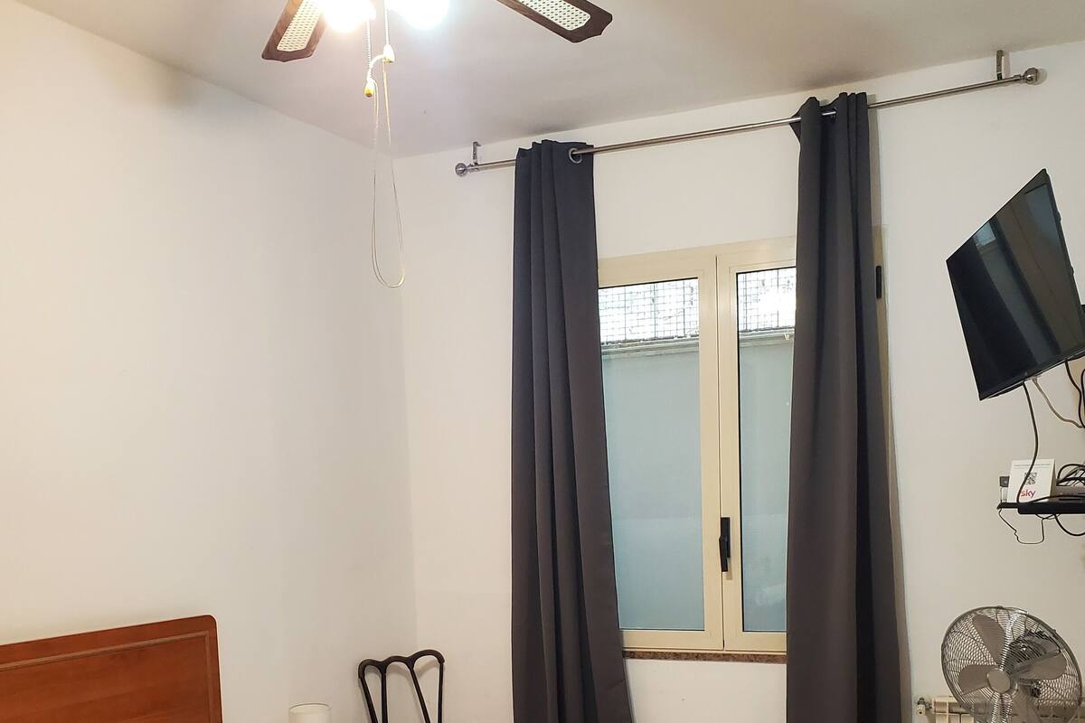 A bedroom with a bed, a nightstand, a chair, a ceiling fan, a TV, and a window with curtains.