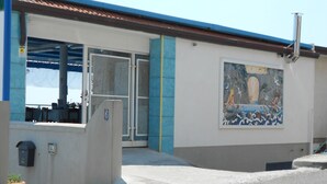 Un edificio con fachada azul y blanca, un mural de una mujer junto al mar, y una entrada protegida por una verja.