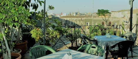 Un patio al aire libre con mesas y sillas, rodeado de plantas en macetas y vistas a un paisaje urbano.