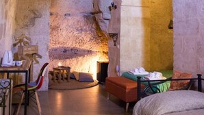 Una habitación acogedora de estilo rústico con techo de piedra, una cama con manta verde y naranja, una pequeña mesa con lámpara, y una silla.