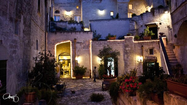 Un antiguo patio de piedra con arcos, plantas en macetas y una escalera que lleva a un nivel superior.