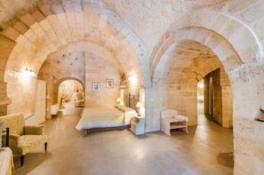 A spacious room with a large bed, a chair, and a desk. The room has stone walls and arches. - Residenza le Dodici Lune (Matera)