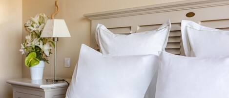 Una cama bien hecha con ropa blanca y almohadas, una mesita de noche con lámpara y un jarrón de flores, y una pared con un diseño sencillo y elegante.
