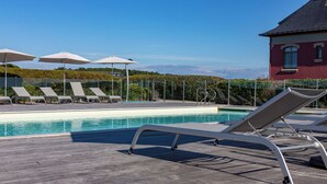 Un área de piscina con sillones de descanso y sombrillas, una terraza de madera y un edificio al fondo.