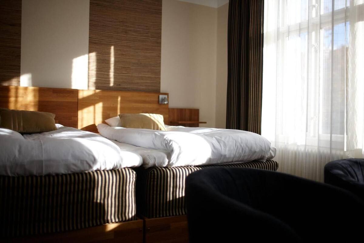 A hotel room with a bed, a wooden headboard, a window with sheer curtains, and a chair.