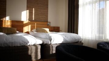 Una habitación de hotel con una cama, cabecera de madera, ventana con cortinas traslúcidas y una silla.