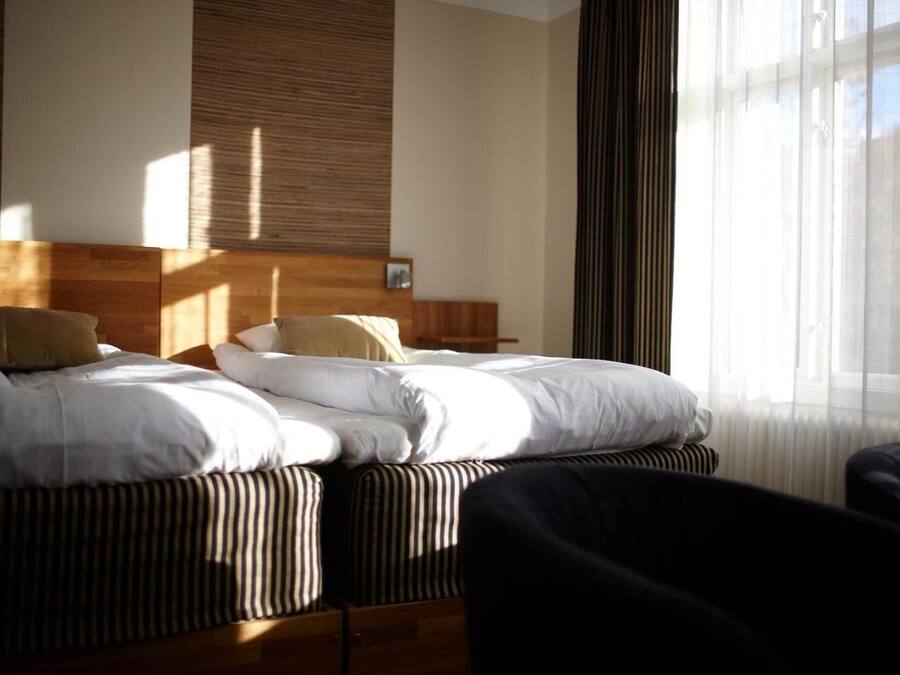 A hotel room with a bed, a wooden headboard, a window with sheer curtains, and a chair.