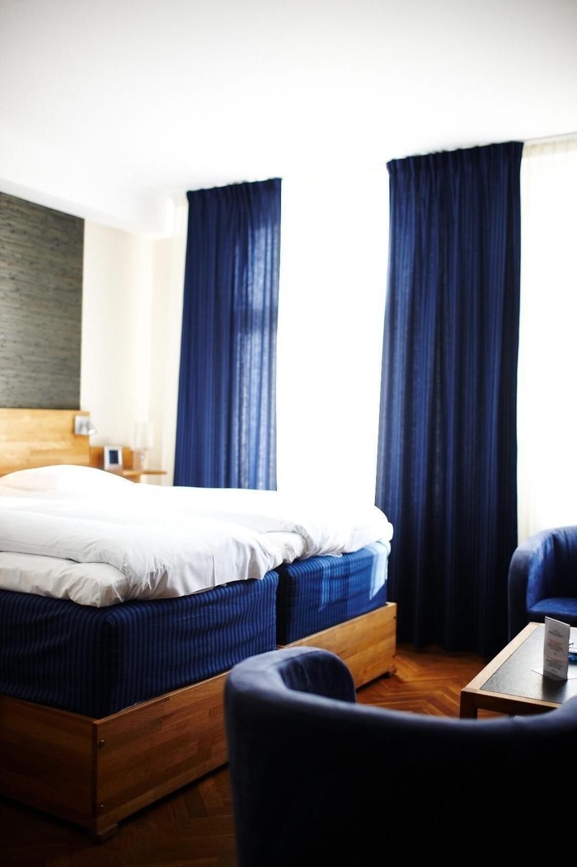 A hotel room with a wooden bed, dark curtains, a wooden nightstand, and a small table with a book.