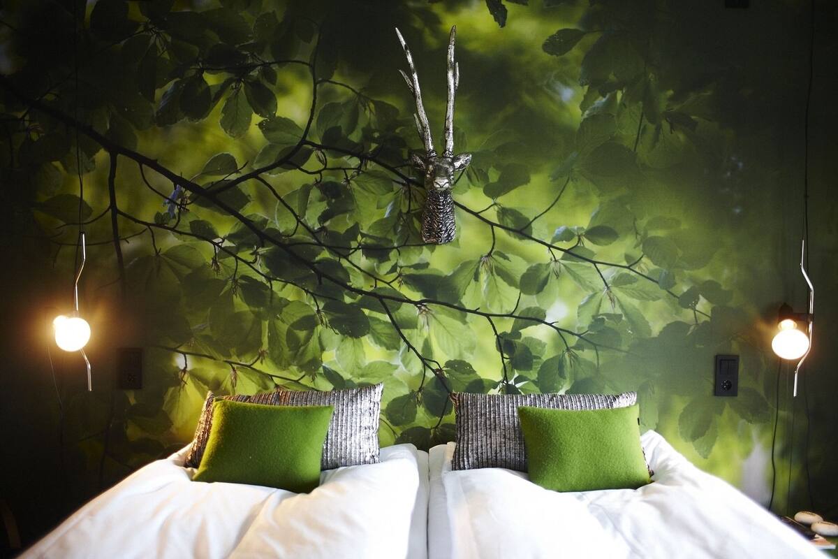 A bed with white bedding and green pillows, set against a wall with a leafy pattern and hanging branches.