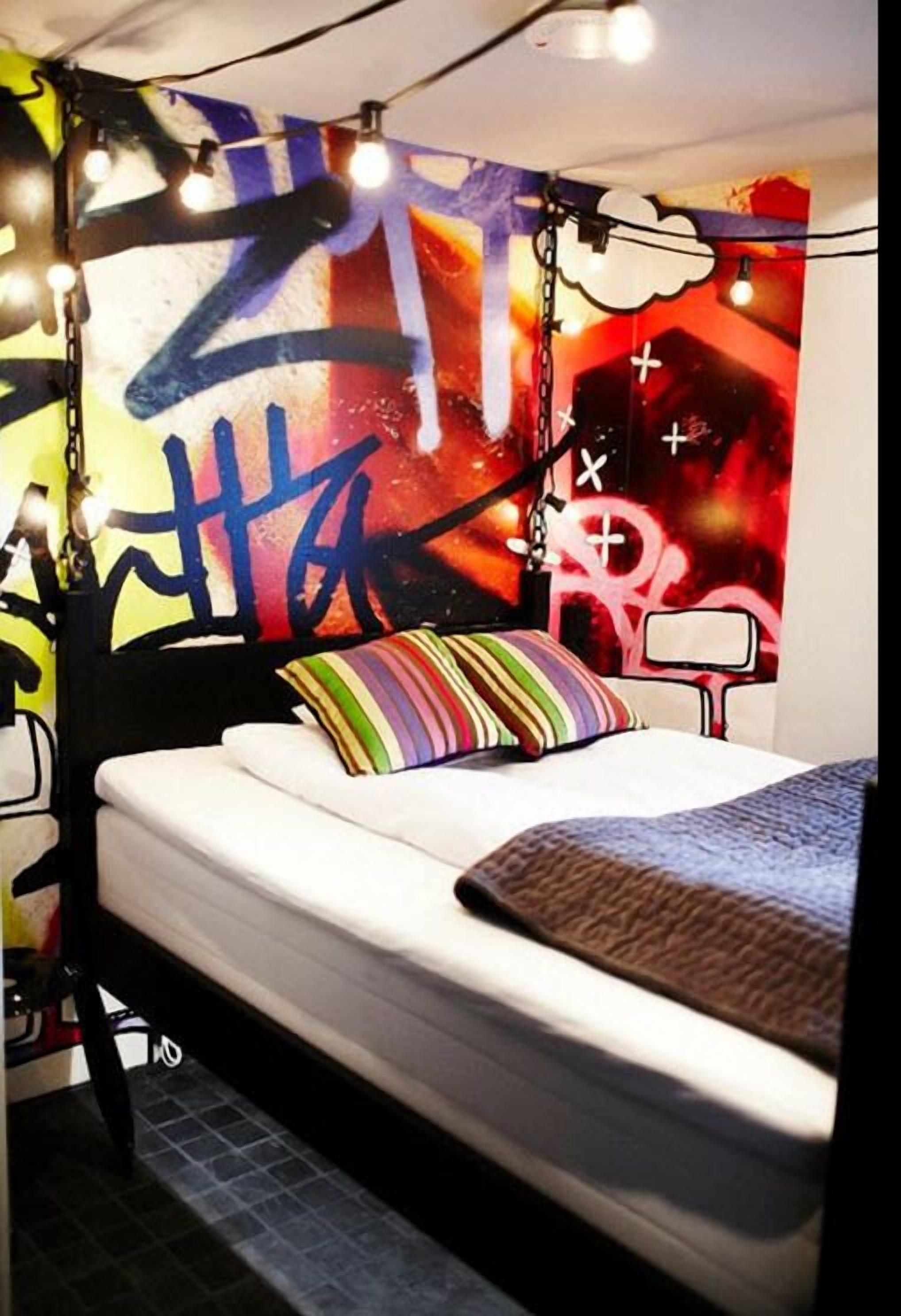 Un dormitorio con una cama, almohadas coloridas, un mural de estilo graffiti y luces colgantes.