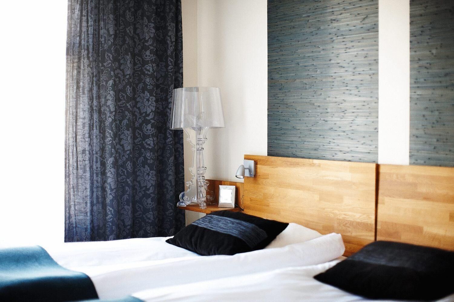 A hotel room with a wooden headboard, a bedside table with a lamp, and a large window with curtains.