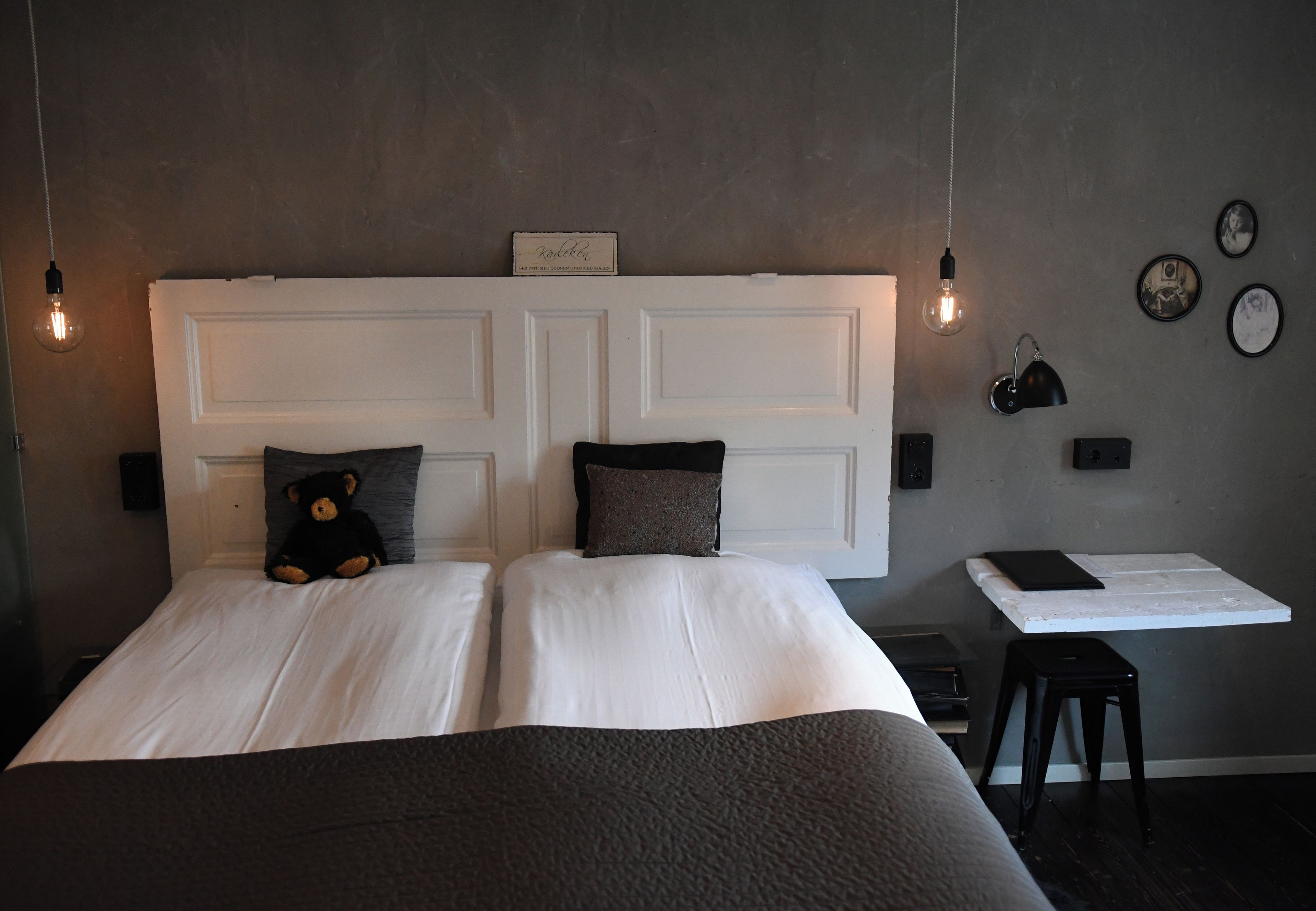 Un dormitorio con una cama grande, dos almohadas, un osito de peluche, una mesita de noche y luces fijadas a la pared.