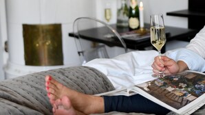 A person lying on a bed with a book, holding a glass of wine, and a vase with flowers in the background.