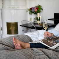 A person lying on a bed with a book, holding a glass of wine, and a vase with flowers in the background.