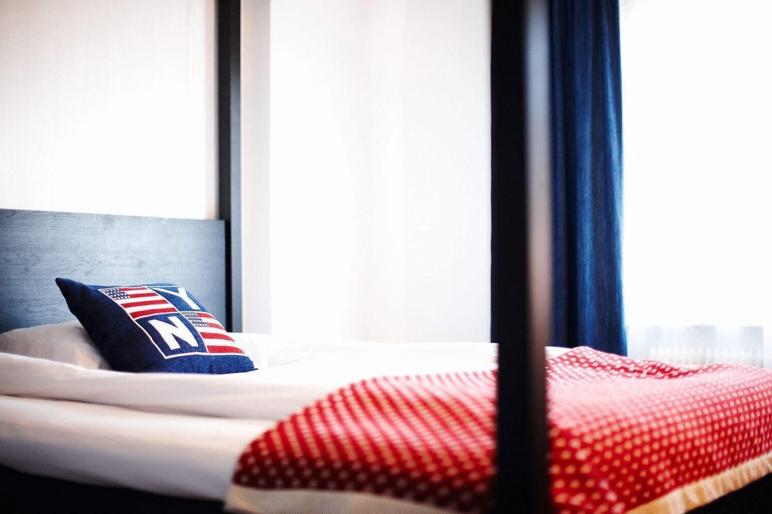 Una habitación de hotel con una cama, un almohadón con diseño de la bandera estadounidense y una ventana con cortinas.