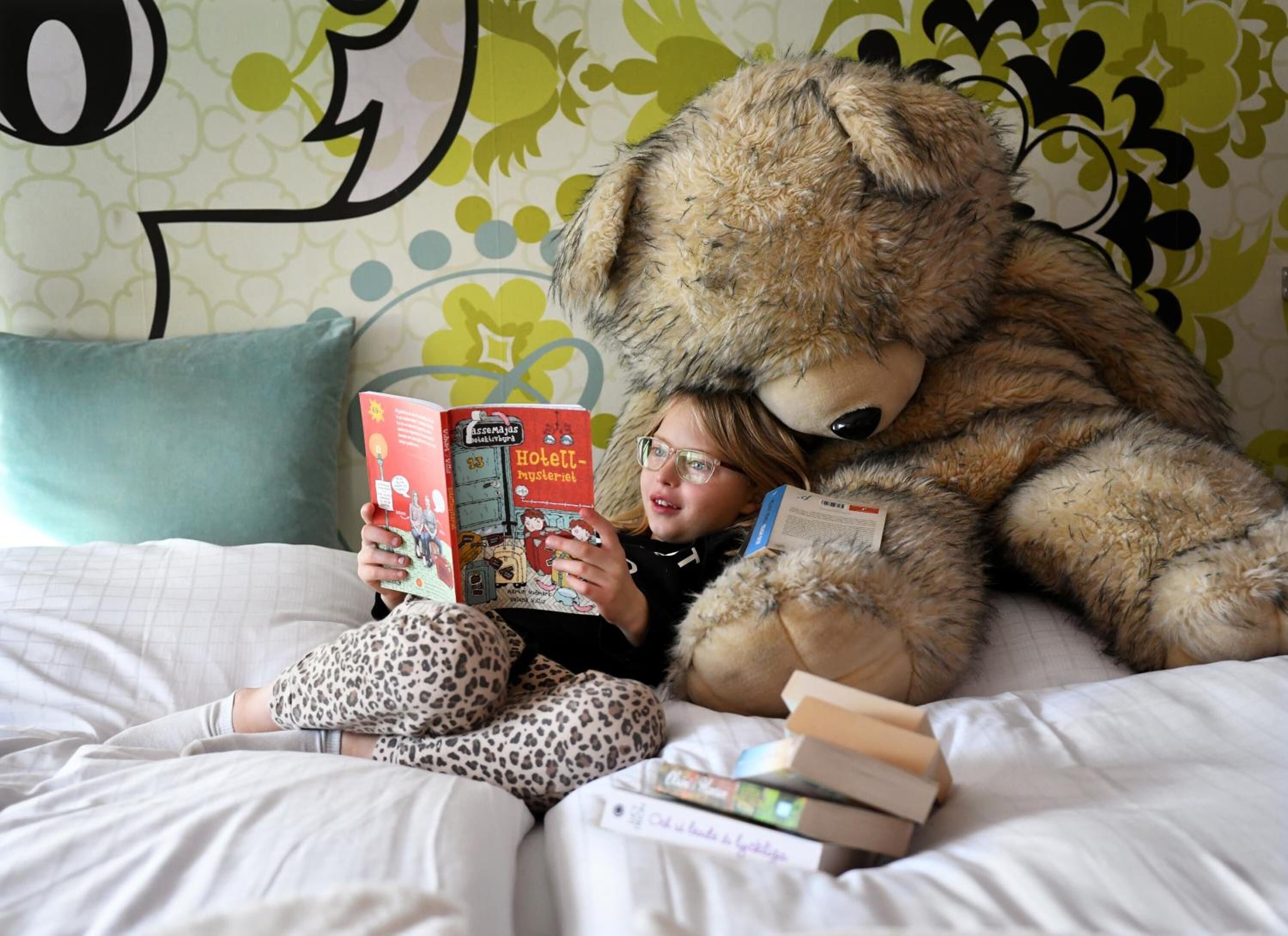Una persona sentada en una cama con un oso de peluche grande y leyendo un libro.