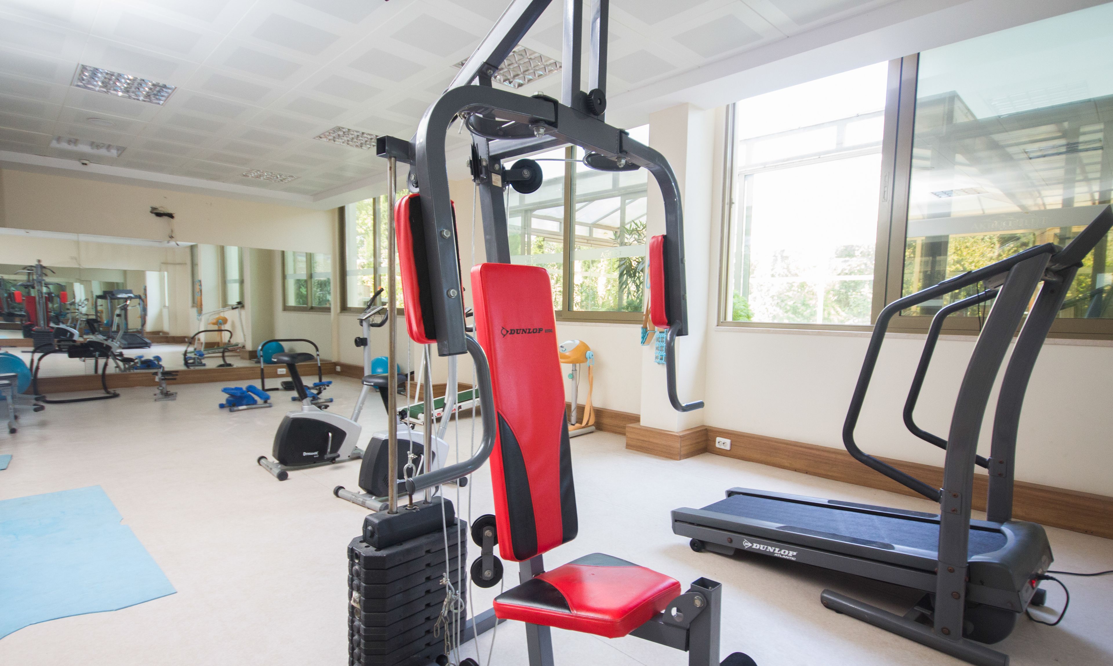 Uma academia com equipamentos de ginástica, esteira e janelas amplas.
