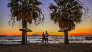 A couple standing under palm trees on a beach at sunset with a view of the sea and distant mountains.