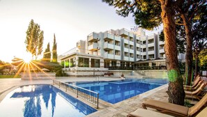 A swimming pool with lounge chairs, a building with balconies, and a tree in the foreground. - Hotel Akbulut (Kusadasi)