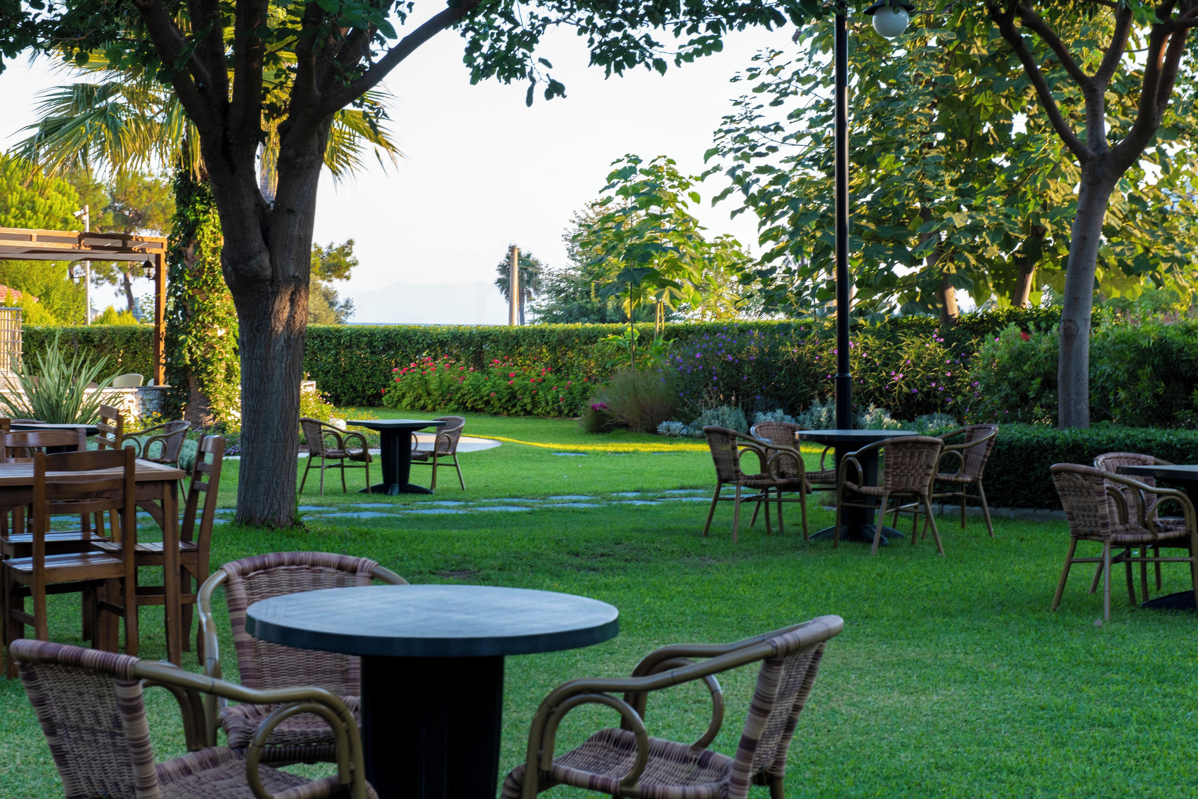 An outdoor patio with wicker furniture and a round table, surrounded by well-maintained hedges and trees.