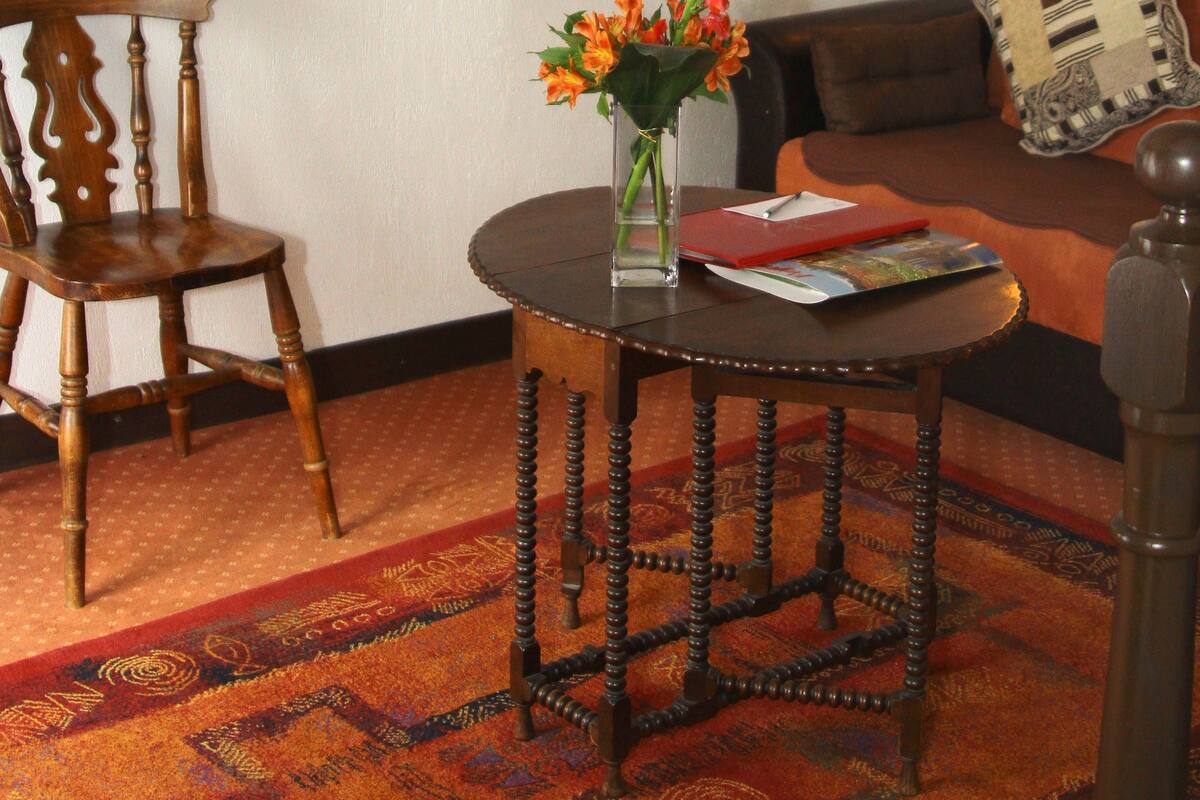 Um quarto com uma cadeira de madeira, uma pequena mesa redonda com um vaso de flores, um quadro na parede, um sofá com uma almofada estampada e um abajur na parede.