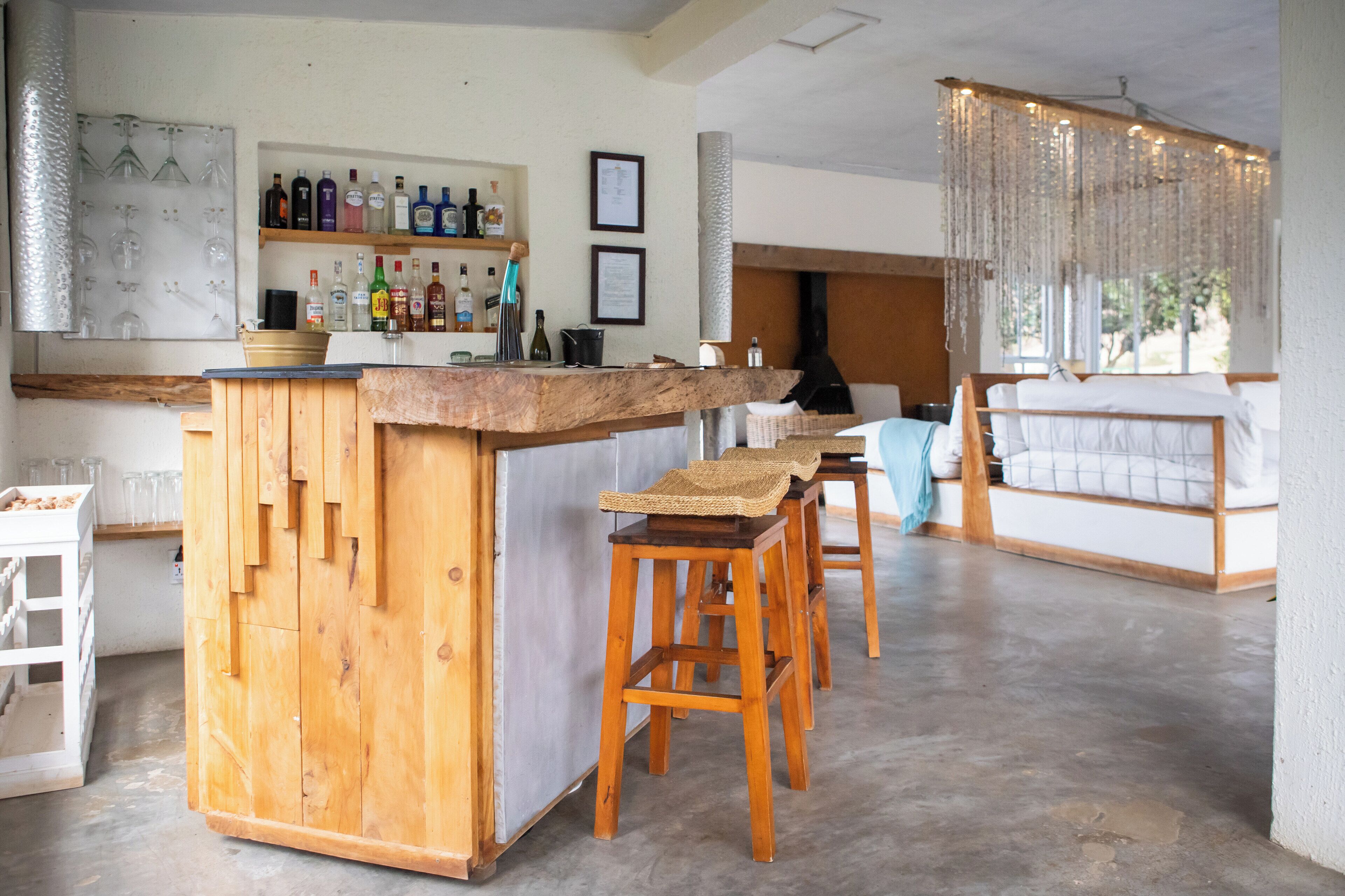 Een bar met houten krukken, een bargedeelte en planken vol flessen. Achtergrond is een bank en een raam met gordijnen.