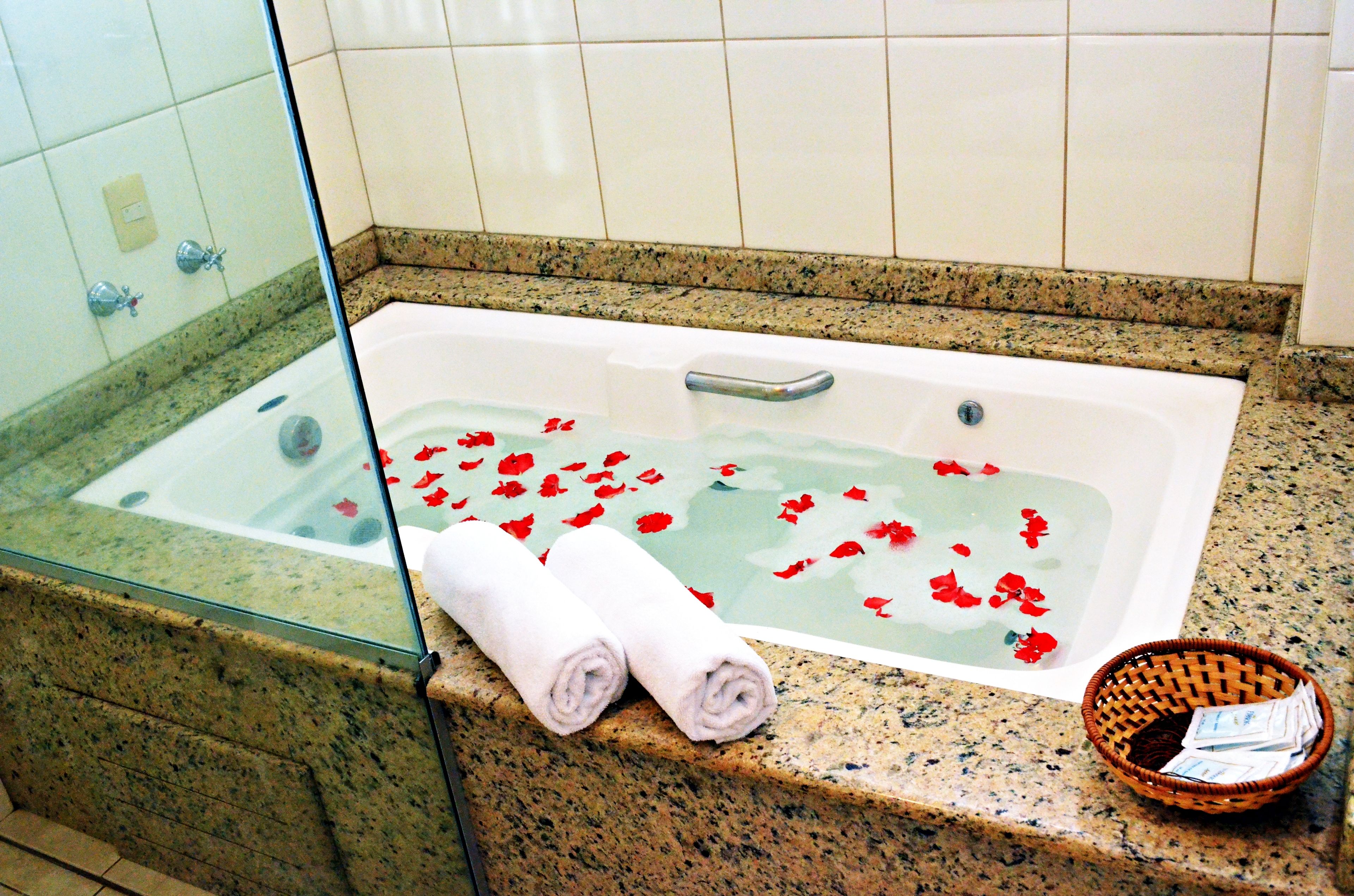 A bathroom with a glass-enclosed bathtub filled with water and red rose petals, two rolled towels on the counter, and a wicker basket with toiletries.